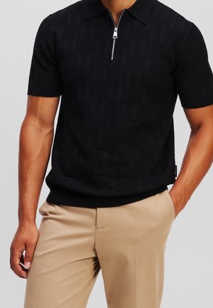 Hombre vestido con polo negro de manga corta con cremallera y pantalones beige, con una mano en el bolsillo, contra un fondo liso.