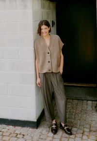 Beige gestrickte ärmellose Strickjacke mit Knöpfen, kombiniert mit grauen hochgeschnittenen Hosen und schwarzen Slippern, vor einer strukturierten Wand.