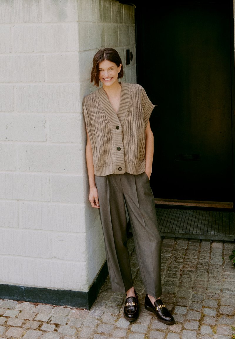 Beige gestrickte ärmellose Strickjacke mit Knöpfen, kombiniert mit grauen hochgeschnittenen Hosen und schwarzen Slippern, vor einer strukturierten Wand.