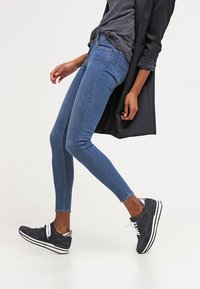 Osoba v úzkých modrých džínech, černém blejzru, pruhované košili a černých teniskách s bílou podrážkou, pózující s mírně pokrčenou nohou.