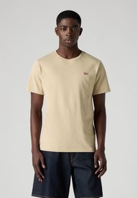 Beige cotton t-shirt with short sleeves and a crew neck, featuring a small red Levi's logo on the chest. Worn with dark denim shorts.