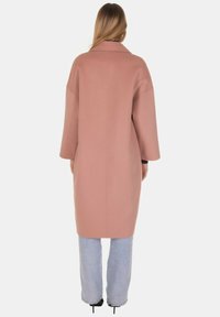 Cappotto lungo oversize in tessuto rosa tenue con colletto con risvolto e maniche larghe, abbinato a jeans azzurri e scarpe nere.