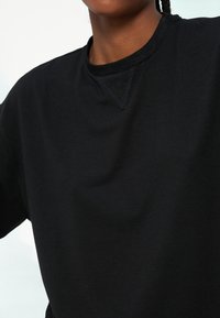 Gros plan d'une personne portant un sweat-shirt noir uni à col rond, montrant l'encolure et la partie supérieure de la poitrine.