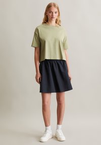 Gesneden olijfgroene T-shirt met korte mouwen, gecombineerd met een marineblauwe gerimpelde rok, afgewerkt met witte sneakers en enkelkousen.