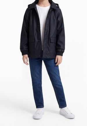 Personne portant une veste à capuche noire sur une chemise blanche, un jean bleu et des baskets blanches, debout devant un fond blanc.