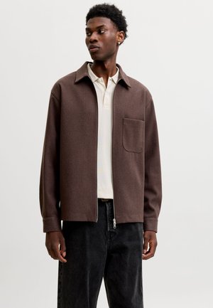Jeune homme portant une veste marron zippée sur un polo blanc et un pantalon noir, regardant vers sa gauche sur un fond uni.