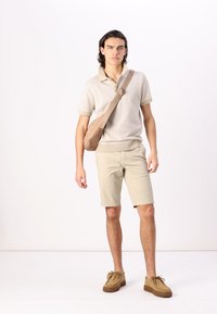 Lys beige polo-skjorte med et teksturert mønster, sammen med beige shorts og brune slip-on sko. En lys brun crossover-veske henger over skulderen.