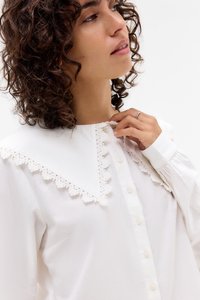 Hvit skjorte med krage som har en scallop-lace kant, knapper foran og lange ermer med knappede mansjetter.
