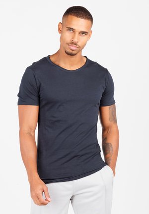 Uomo con capelli corti e tatuaggi, che indossa una maglietta aderente blu scuro e pantaloni chiari, guarda leggermente verso l'alto a destra.