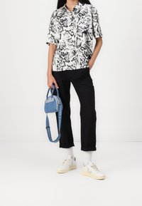 Chemise blanche à manches courtes avec motif floral noir, pantalon noir, baskets ton gris et un petit sac à main bleu avec une longue bandoulière.