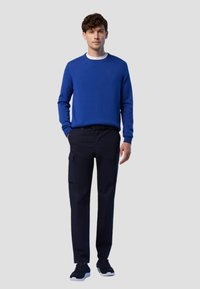 Maglione blu con scollo rotondo, abbinato a pantaloni cargo blu scuro. Il maglione presenta un piccolo emblema, indossato con sneaker nere.