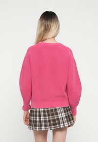 Sweater de malha rosa com textura canelada, gola redonda e mangas balão, combinado com uma saia plissada em padrão xadrez nas tonalidades castanho e branco.