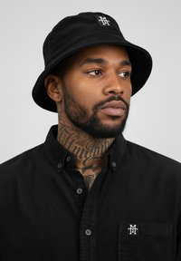 Black bucket hat with a small white logo on the front, worn with a black button-up shirt featuring a chest pocket and classic collar.