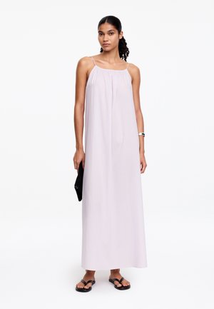 Femme aux cheveux attachés portant une robe longue sans manches rose pâle, des sandales noires, une pochette noire et un bracelet argenté, debout devant un fond blanc.