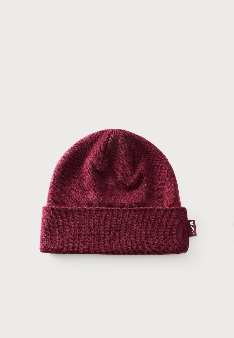 Bonnet en maille bordeaux avec revers plié et petite étiquette blanche portant la mention « GOLF » sur un fond clair uni.