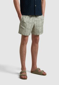Heren shorts in lichtgroen met een wit golfpatroon, voorzien van zijzakken. Gdragend met grijze slipper sandalen. Aangepaste tailleband.