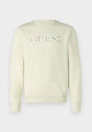 Kerge beeži värvi ringkaelusega sviiter, millel on valge "GUESS EST. 1981" tikand rinnal, pikad varrukad ja soonikdetailid.