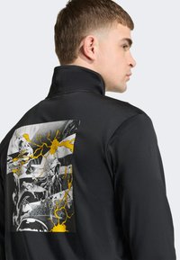 Czarna kurtka z wysokim kołnierzem posiada na plecach grafikę łączącą elementy czerni, bieli i żółci, prezentując dynamiczne ilustracje.