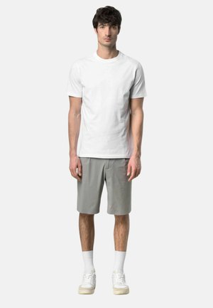 Giovane uomo con capelli scuri in piedi dritto, che indossa una semplice t-shirt bianca, pantaloncini grigi fino al ginocchio, calzini bianchi e scarpe da ginnastica bianche.