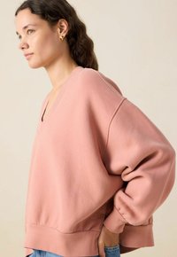 Sweatshirt rose clair à col en V avec des épaules tombantes, des poignets et un ourlet côtelés. Le tissu semble doux, avec des coutures minimales et une coupe décontractée.