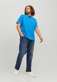 Polo azul con collar a rayas en blanco y negro; combinado con jeans oscuros y zapatillas blancas. El modelo se encuentra de manera casual, con las manos en los bolsillos.