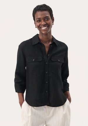 Camicia nera in lino con due tasche al petto, colletto e maniche lunghe. Indossata con pantaloni bianchi a righe.