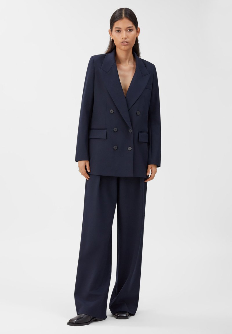 Eine Frau steht in einem marineblauen, doppelreißigen Oversized-Blazer mit weit geschnittenen passenden Hosen und schwarzen Rechteckzehen-Schuhen.