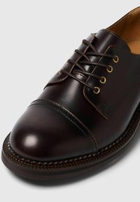 Scarpa in pelle marrone scuro con punta tondeggiante, design classico con lacci, occhielli dorati e cuciture a contrasto lungo la suola.