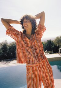 Femme souriante aux cheveux bouclés, portant un ensemble assorti orange et blanc à motifs, debout près d'une piscine avec des arbres en arrière-plan.