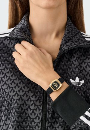 Must zip-up jakk musta-halli lehemustriga ja valgete triipudega, millel on kuldse ja musta ruudukujuline kell mustal rihmal.