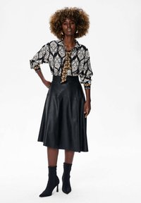 Vrouw met krullend haar draagt een gemusterde blouse met lange mouwen, een zwarte leren midi rok, een gemusterde stropdas en zwarte haklaarzen, staande tegen een witte achtergrond.