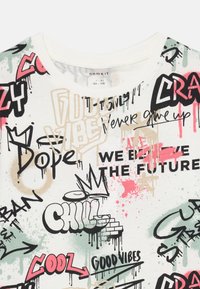 Baumwoll-T-Shirt mit rundem Halsausschnitt, das bunte Graffiti-ähnliche Drucke in Schwarz, Pink, Grün und Beige auf einem cremefarbenen Hintergrund zeigt.