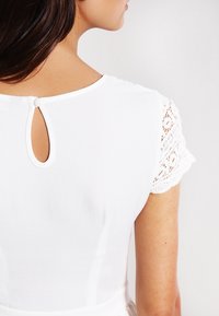 Mujer vistiendo un vestido blanco con mangas cortas de encaje y un pequeño cierre de botón en forma de lágrima en la parte posterior del cuello.