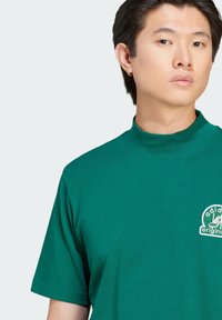 Green cotton t-shirt featuring a high neckline and short sleeves. It has an embroidered logo in white on the left chest.