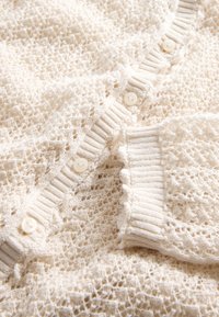 Strickjacke in Cremefarbe, gefertigt aus einem strukturierten, offenen Gewebe, mit gerippten Kanten und leichten, runden Knöpfen.