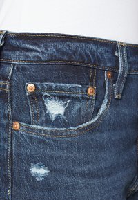 Jeans de denim con un lavado oscuro, que presentan un bolsillo desgastado con bordes deshilachados, remaches de bronce y costuras amarillas en contraste.