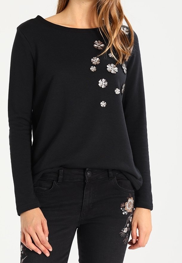 Svart långärmad topp med broderade blommönster i olika färger på vänster sida, som matchas med svartajeans med blommiga detaljer.