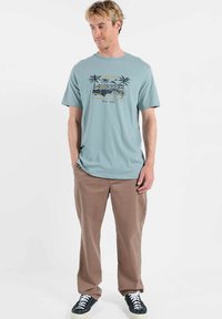 T-shirt en coton bleu clair avec un motif graphique de palmiers et un véhicule, associé à un pantalon marron clair et des baskets foncées.