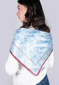 Écharpe en soie à motifs bleus et blancs avec un design floral, ornée d'une bordure en satin bordeaux, portée sur l'épaule.