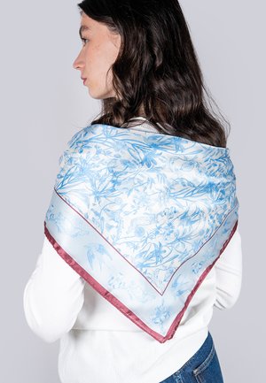 Écharpe en soie à motifs bleus et blancs avec un design floral, ornée d'une bordure en satin bordeaux, portée sur l'épaule.
