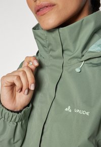 Grønn Vaude-jakke har høy krage, teksturert stoff, justerbar snor og brodert logo. Håndtakene har elastisk strikker.