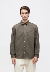 Joven con una camisa verde oliva abotonada y bolsillo en el pecho, y pantalones crema, de pie contra un fondo claro liso.