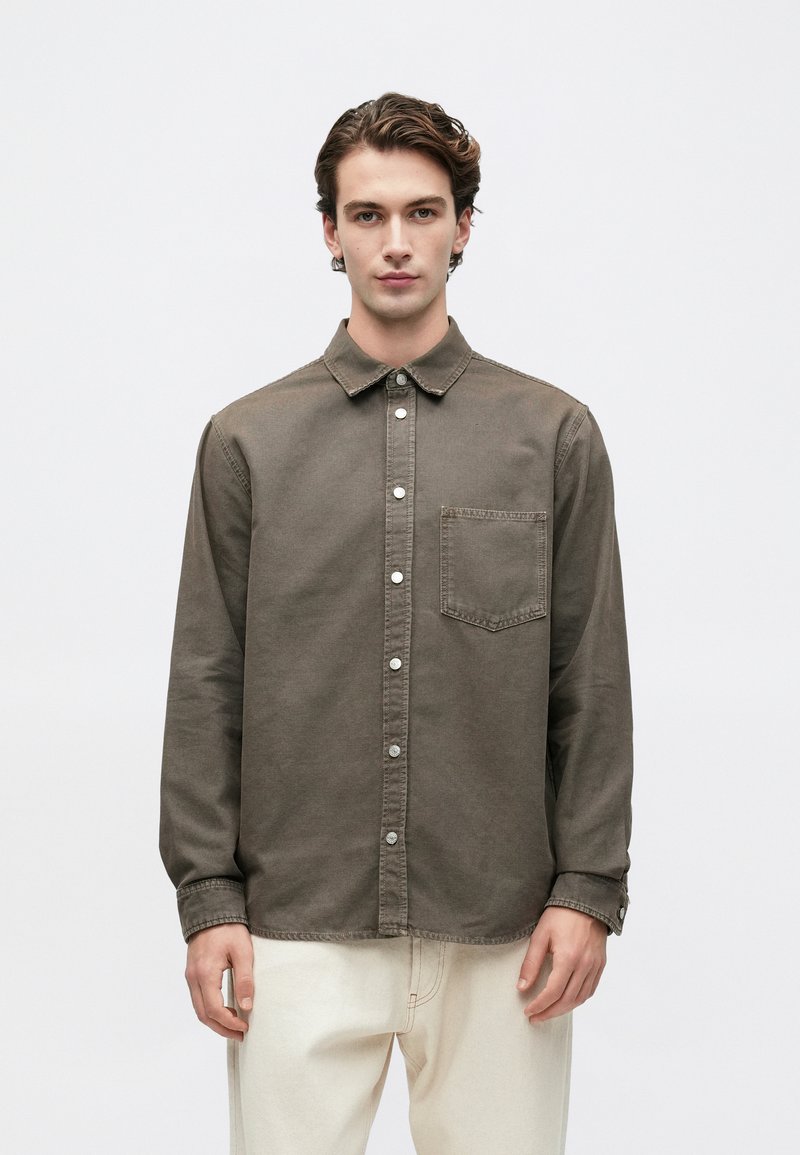 Joven con una camisa verde oliva abotonada y bolsillo en el pecho, y pantalones crema, de pie contra un fondo claro liso.