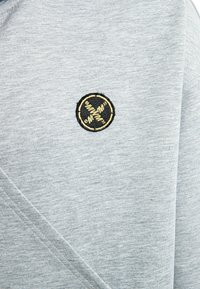 Sudadera gris con una textura suave, diseño cruzado y un logotipo bordado circular en negro y dorado en el pecho.
