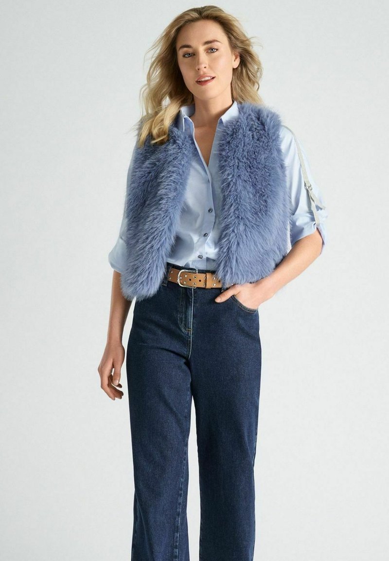 Lichtblauwe nepbont gilet over een lichtblauw overhemd met knopen, gecombineerd met donkerdenim wijde jeans en een lichtbruine riem.