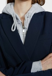 Blazer blu navy sopra una felpa grigia chiara con zip. La felpa ha il cappuccio con stringhe e una consistenza morbida. Il blazer presenta una tasca sul petto.