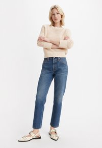 Suéter de lana crema con tejido abierto, jeans azul de cintura alta y zapatos blancos con correas ajustables y acentos de tachuelas.