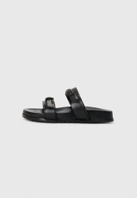 Sorte skinnsandaler med to brede stropper, en med metallspenne, og en flat, teksturert såle med et rillet mønster.