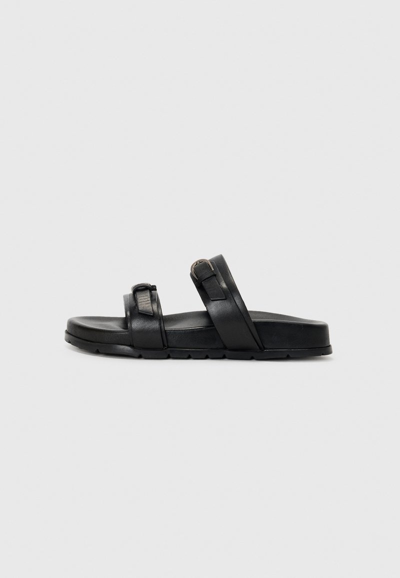Sorte skinnsandaler med to brede stropper, en med metallspenne, og en flat, teksturert såle med et rillet mønster.