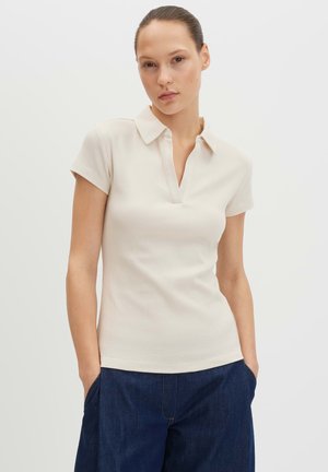 Jeune femme à la peau claire et aux cheveux tirés en arrière, portant un polo crème à manches courtes et un pantalon bleu foncé, les mains dans les poches.